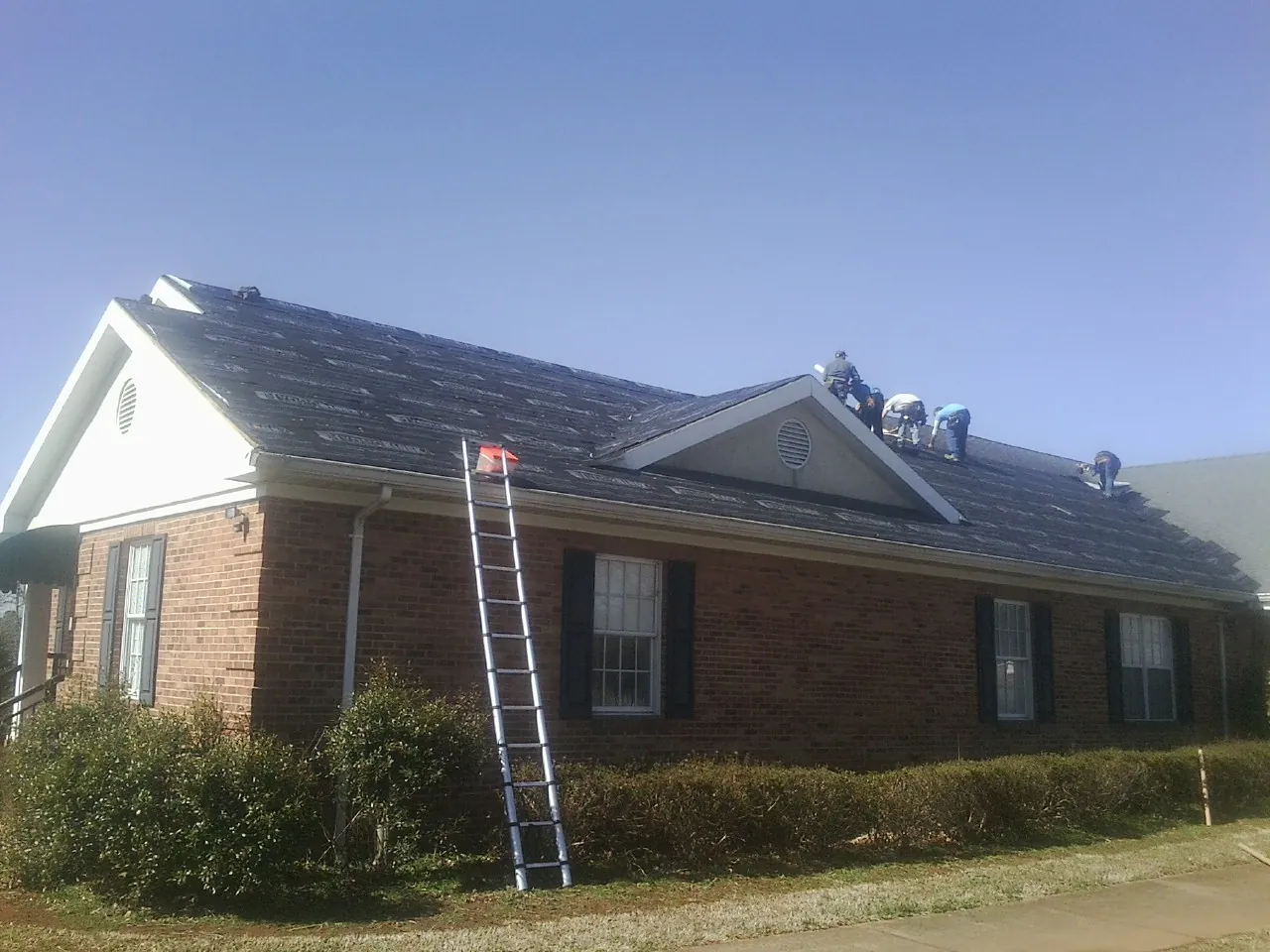 Crew tearing shingles off a brick ranch roof with a ladder set — Emergency Roof Repair work by our crew in Clara City