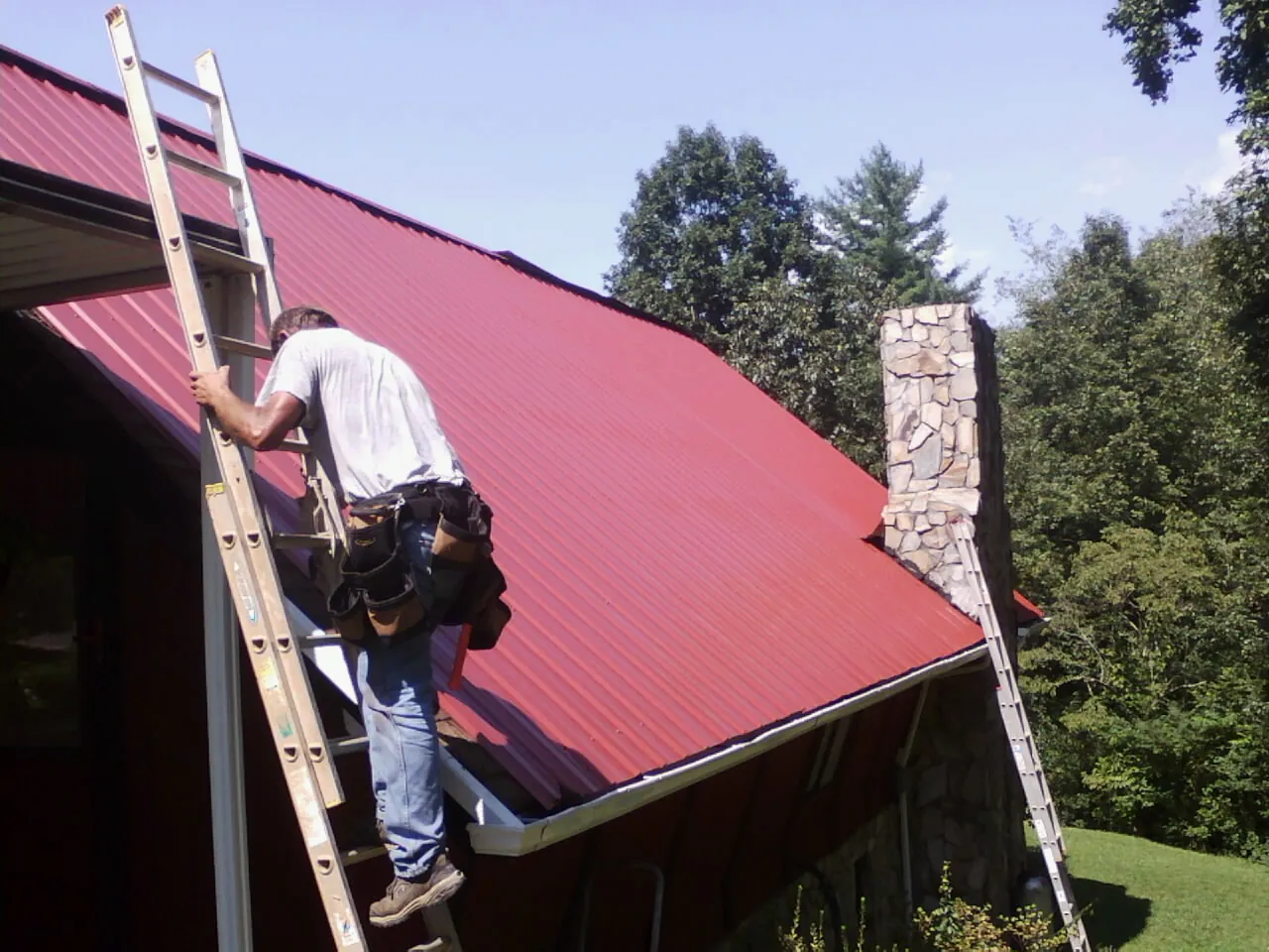 Roofer on a ladder beside a red metal roof and stone chimney — Soffit & Fascia Repair by Roofing Brothers in Clara City